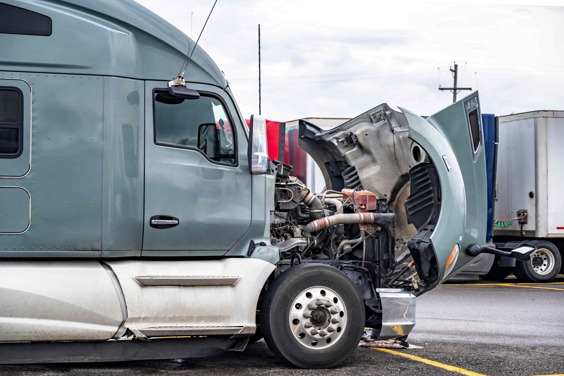 Damaged semi-truck cab after collision, hood crumpled open on a wet highway.