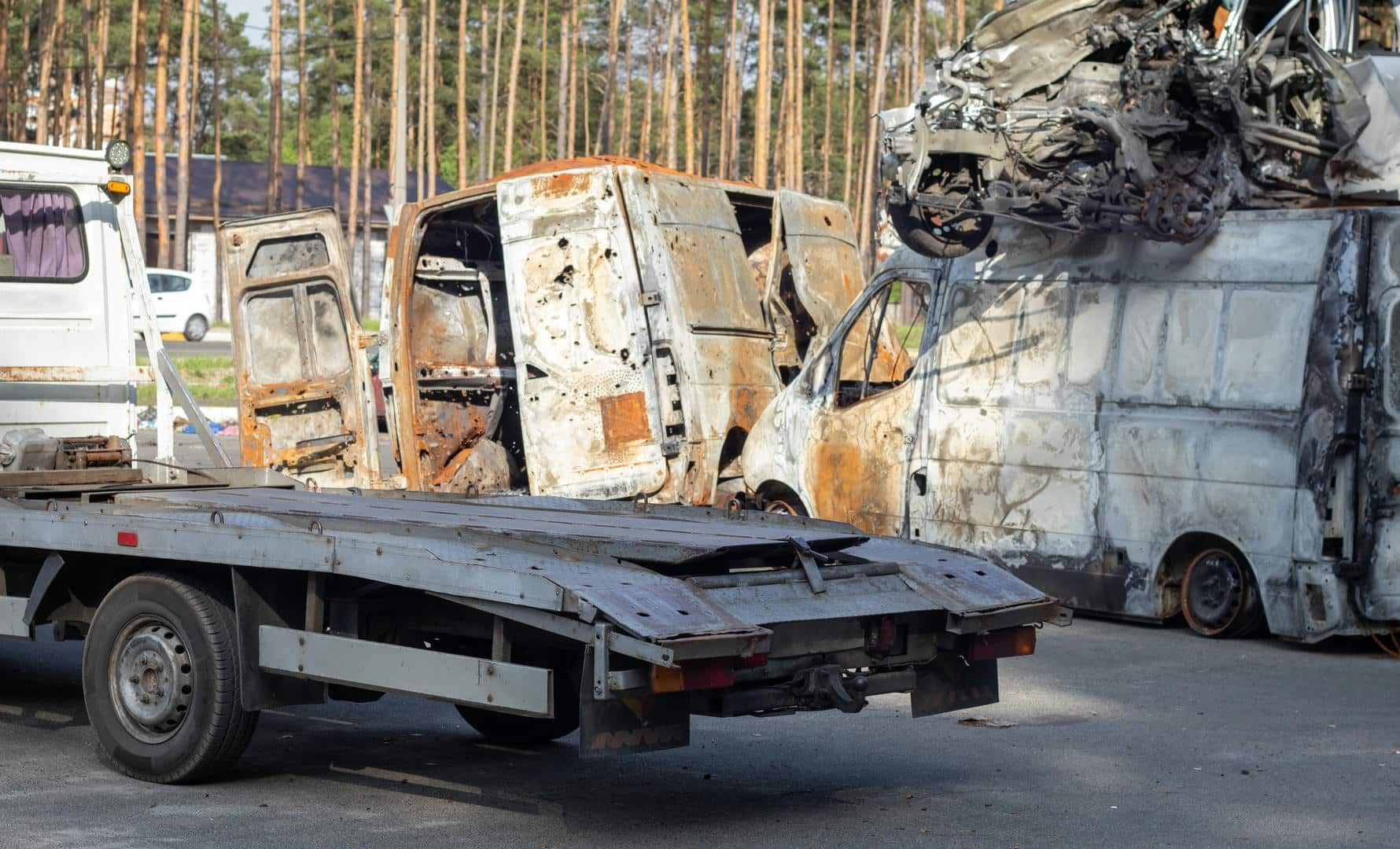 Damaged vehicles on tow truck, likely after a conflict, in a daylight setting. Burn marks and bullet holes are visible.