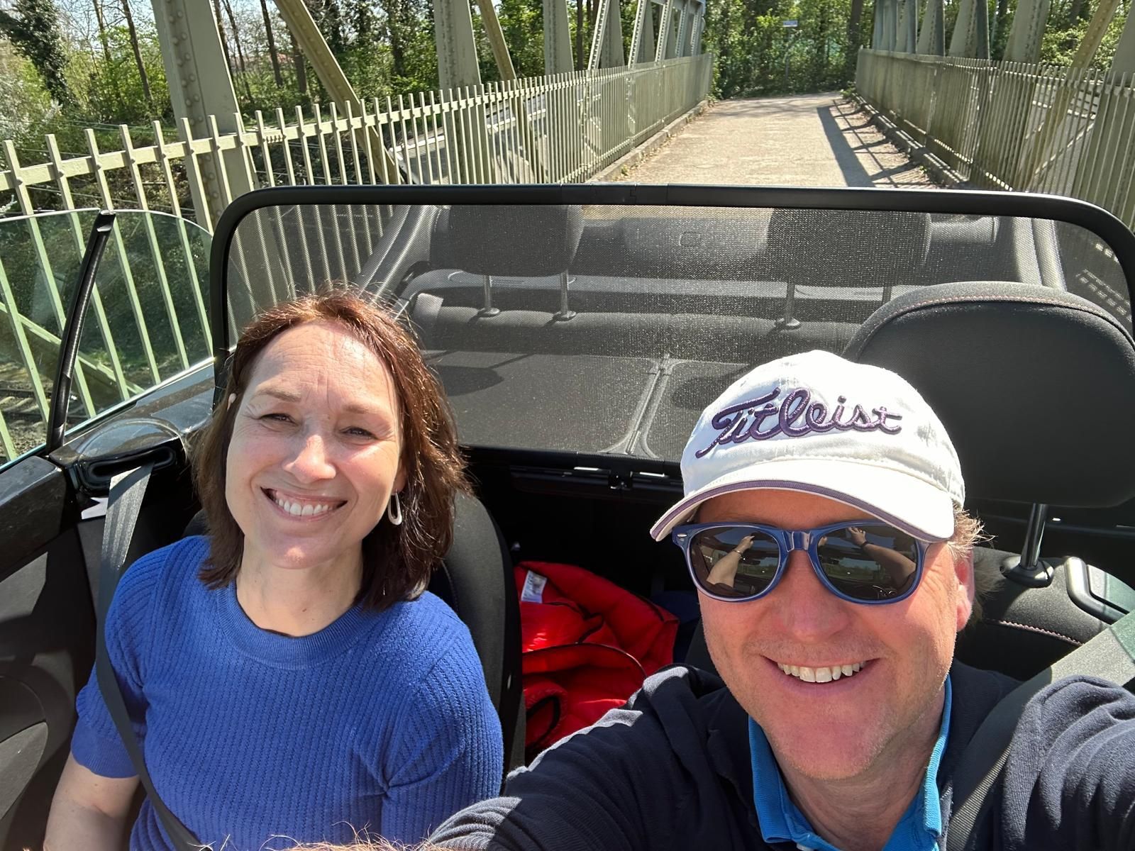 Een lachend stel in een cabriolet rijdt op een brug op een zonnige dag.