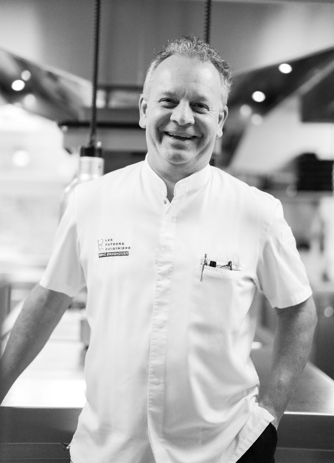 Een chef-kok in wit uniform glimlacht in een professionele keuken, met zijn handen op het aanrecht.