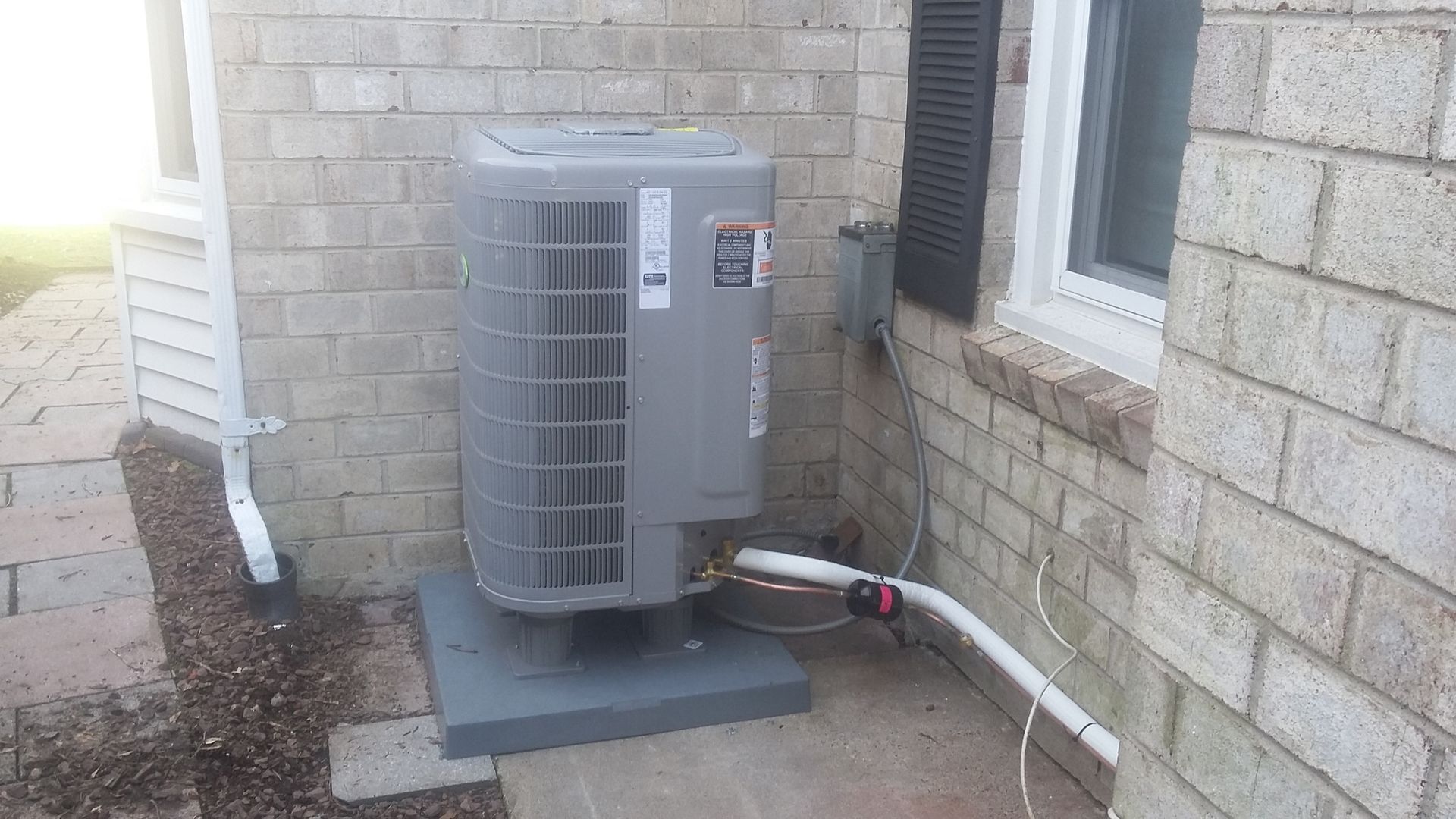 An air conditioner is sitting on the side of a brick building next to a window.