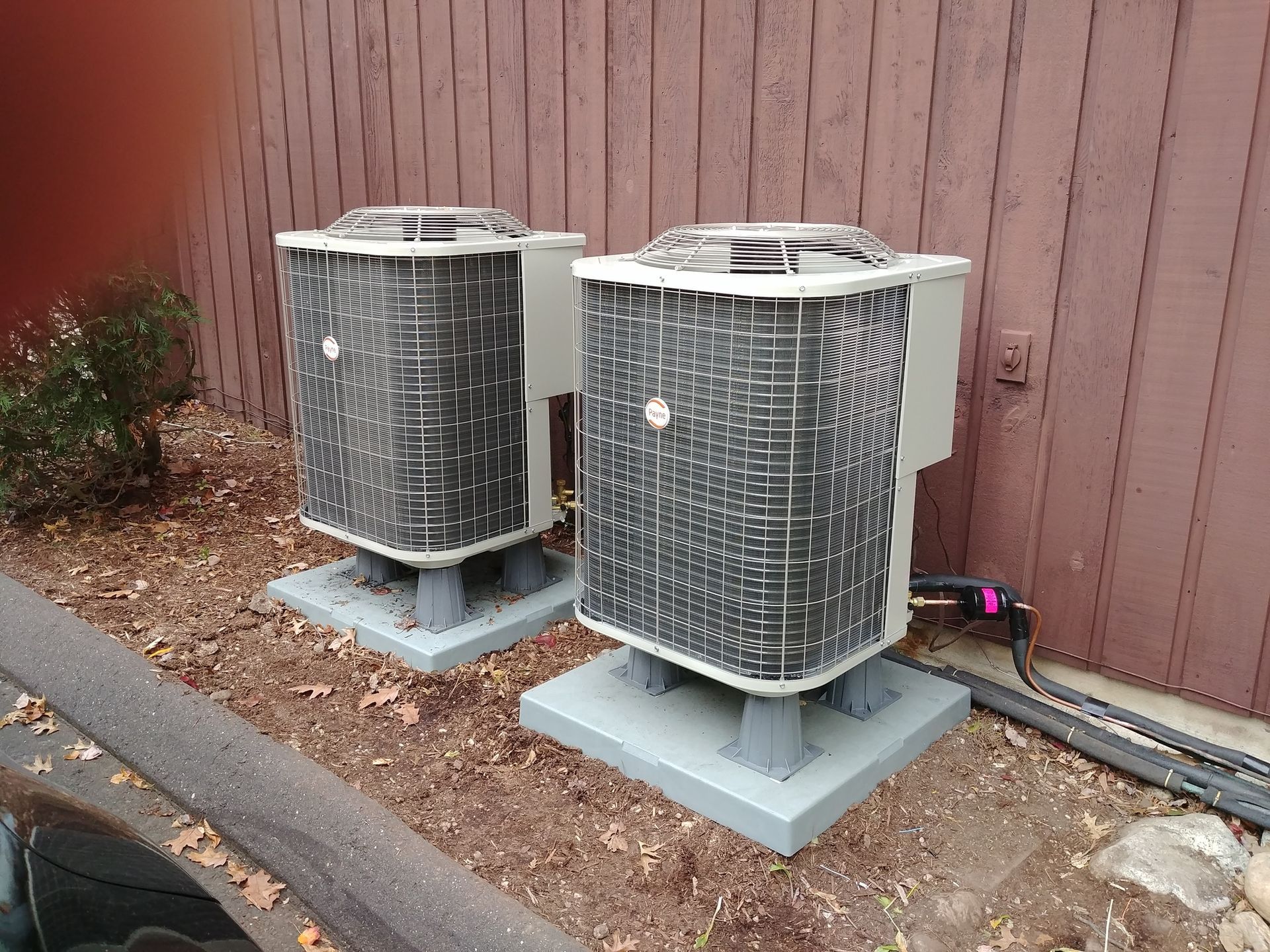 Two air conditioners are sitting next to each other in front of a wooden fence.