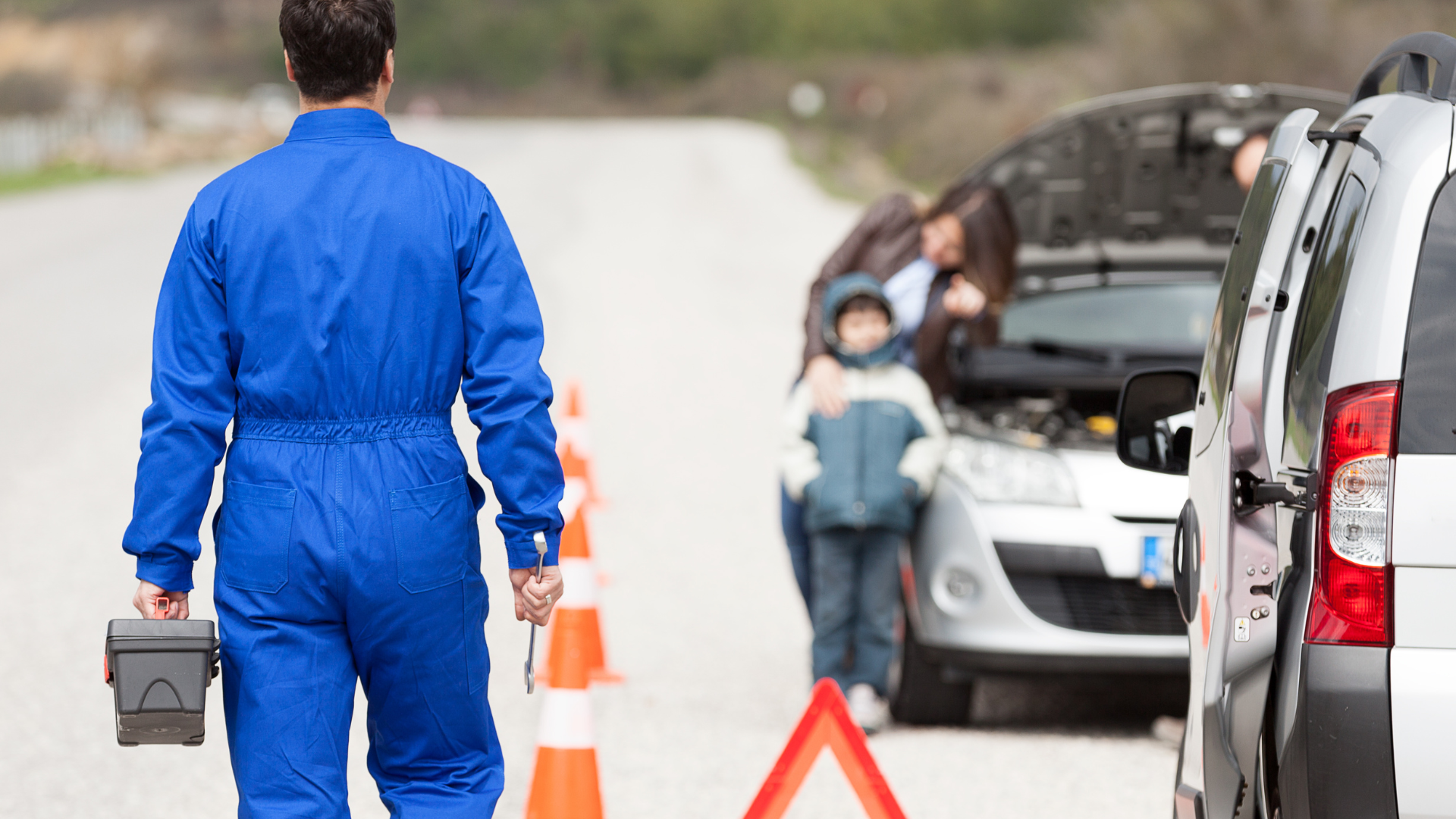 Mechanic in blue overalls walks toward a broken-down car, family looking at open hood.