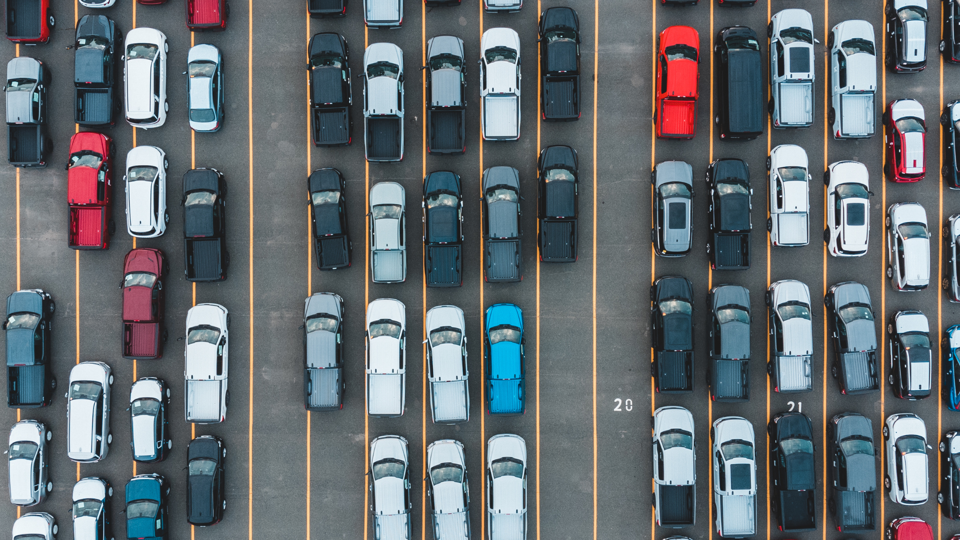 Overhead view of a crowded parking lot filled with various cars, primarily trucks, in shades of white, gray, and black.