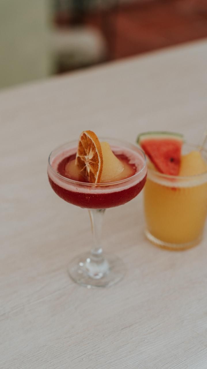 Dos cócteles coloridos sobre una mesa, uno rojo con una rodaja de naranja y otro naranja con una guarnición de pomelo.