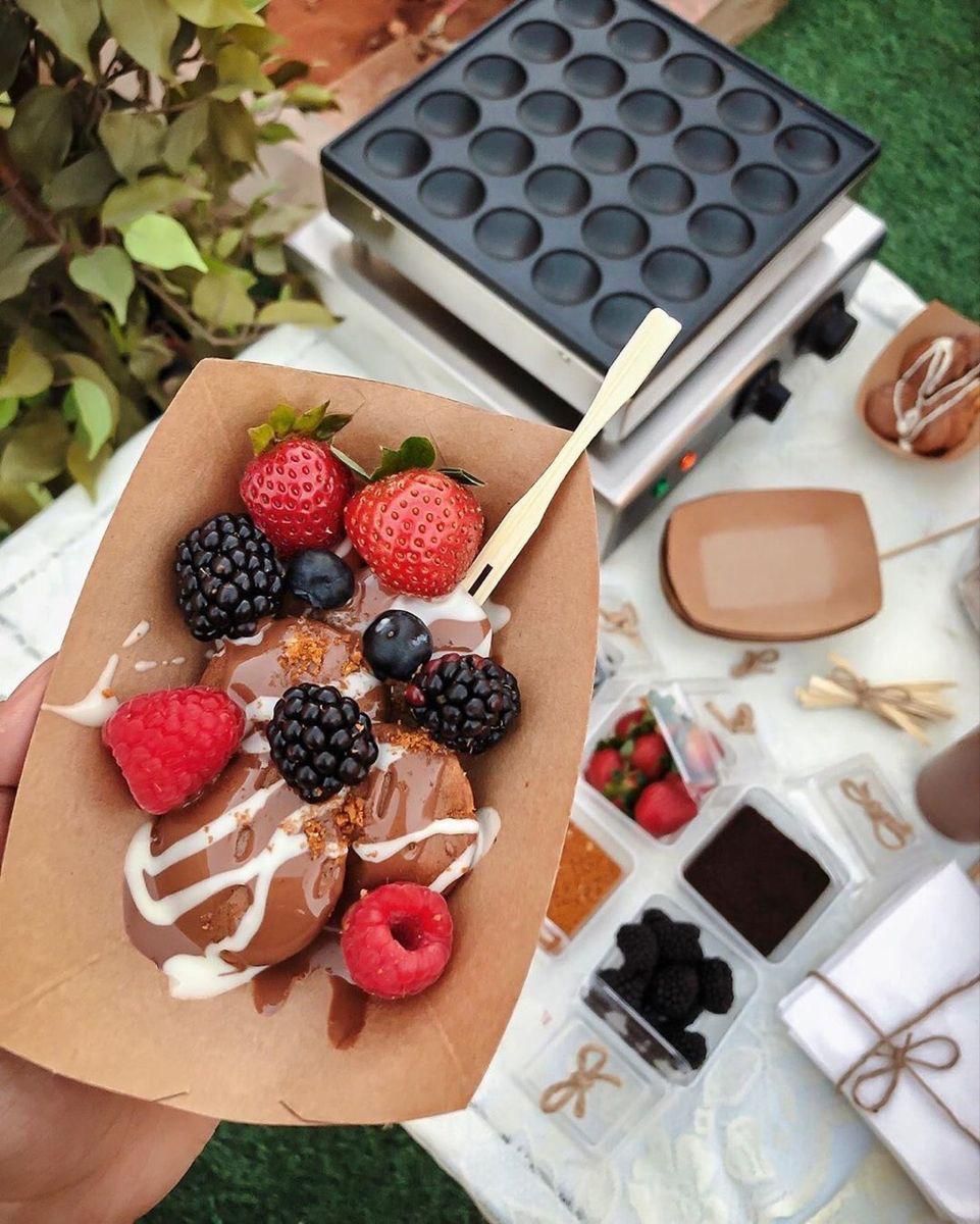 Cono de papel con gofres cubiertos de frutos rojos y glaseado blanco, junto a una mini gofrera y frutos rojos.