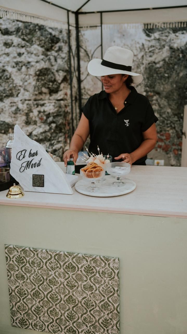 Hombre con camisa negra y sombrero blanco sirviendo bebidas en una mesa de postres decorada al aire libre.