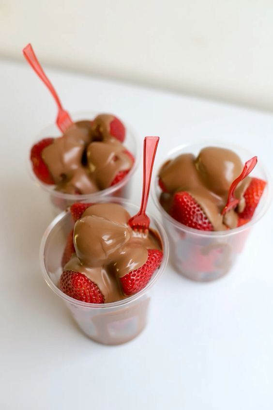 Tres vasos transparentes de postre de fresa y chocolate con cucharas rojas sobre una superficie blanca.
