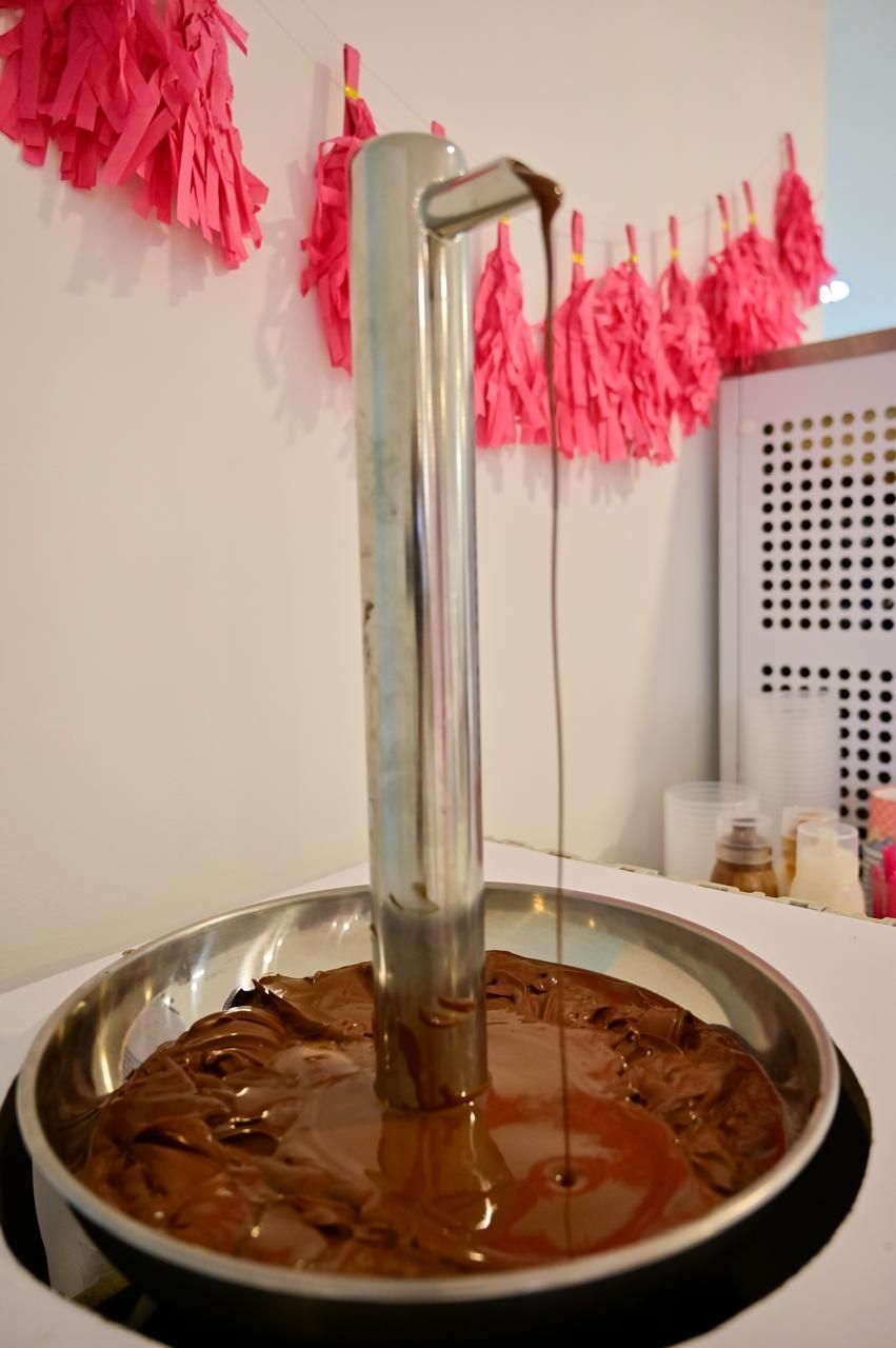Boquilla metálica que dispensa chocolate en un recipiente redondo de metal, con borlas rosas al fondo.