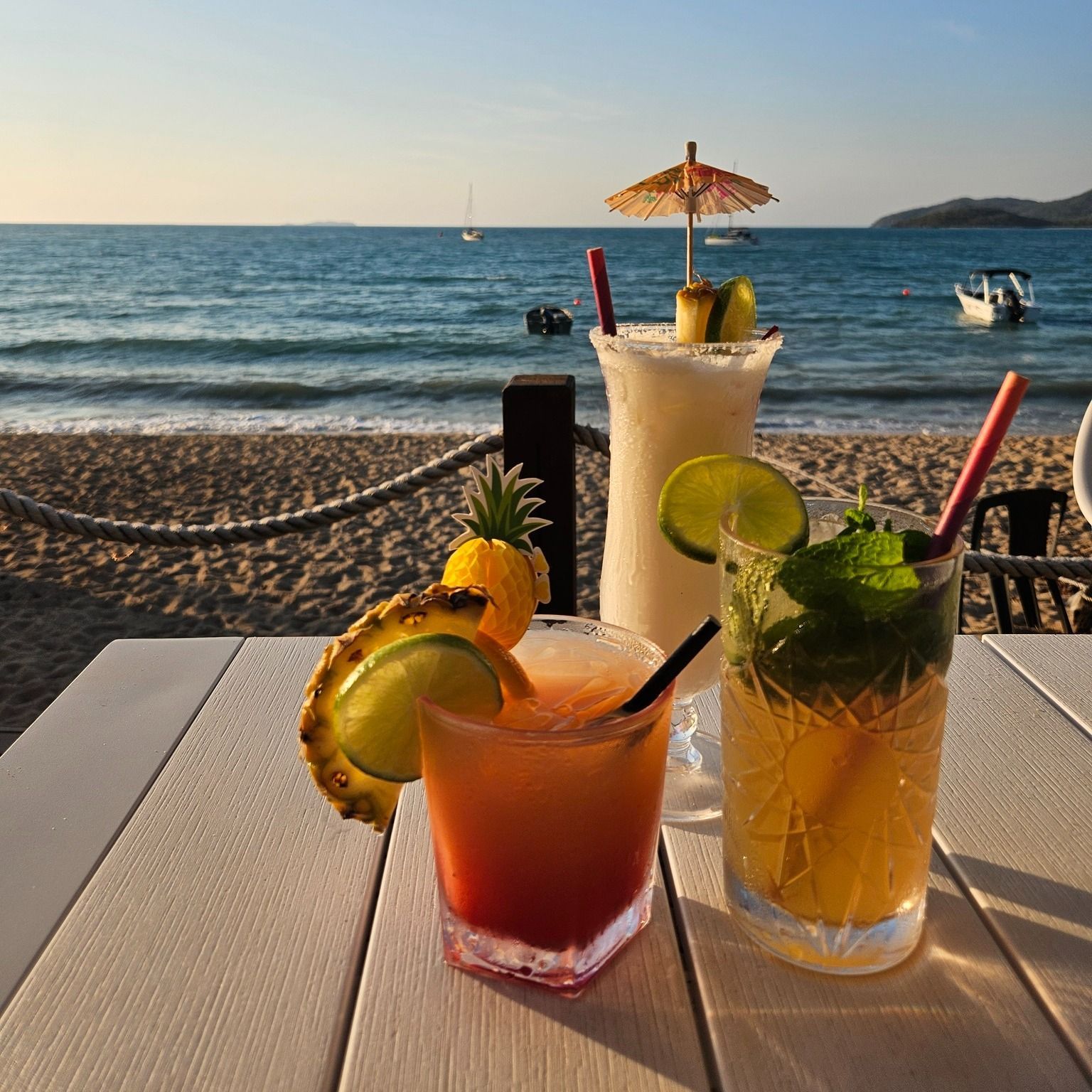 Three tropical cocktails on a table overlooking a beach — The Beach Bar and Restaurant at Montes in Cape Gloucester, QLD