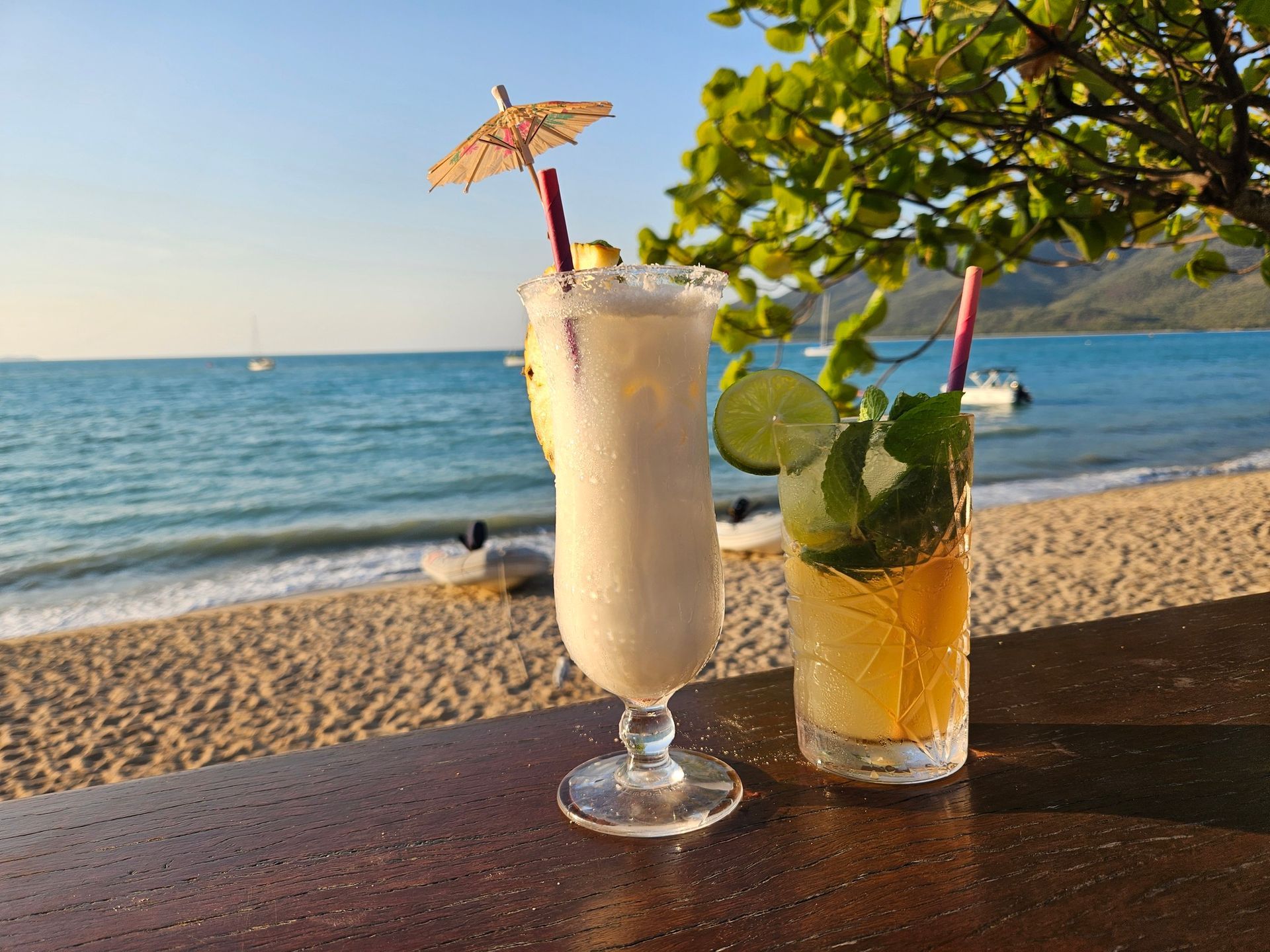 Cocktails on a bar, beach background. A white drink with umbrella, a yellow-green drink, blue ocean, sunny day.