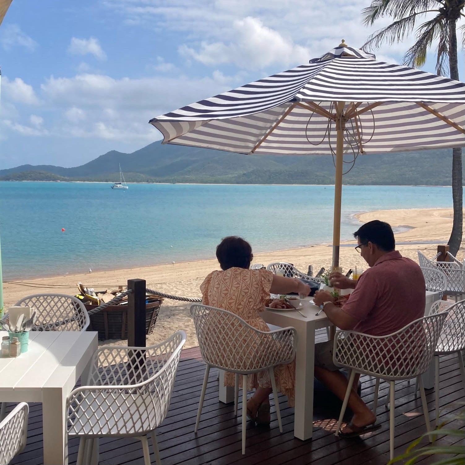 Couple dining at beachside table under a striped umbrella — The Beach Bar and Restaurant at Montes in Cape Gloucester, QLD