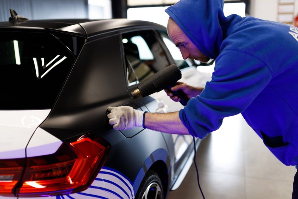 Man Using Heat Gun on Car's Black Vinyl Wrap — DXMA Designs In Narellan, NSW