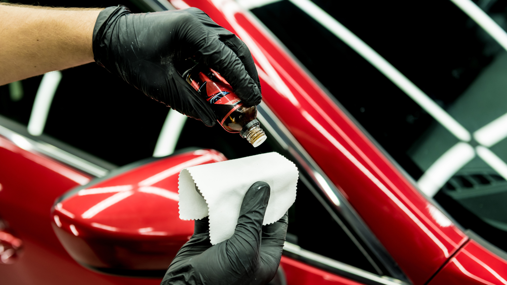 Person with black gloves applying liquid to a white cloth, red car in the background.