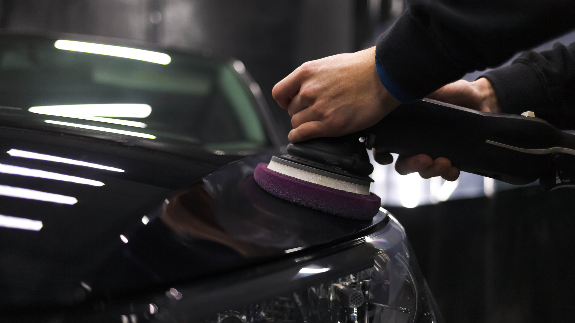 Person polishing a black car with a power buffer, indoors.