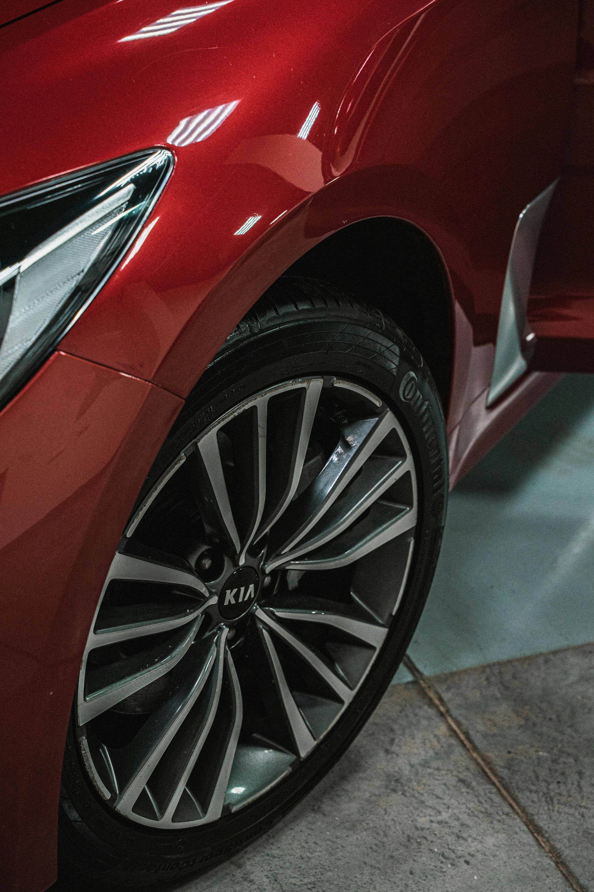 Red Kia car, close-up of front wheel and fender. Silver and black alloy wheel, on gray surface.
