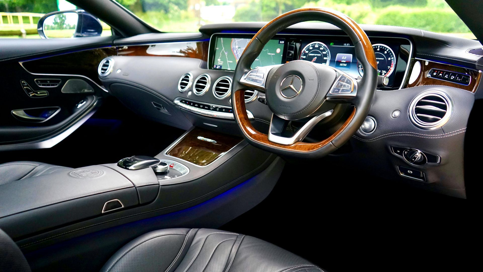 Mercedes-Benz car interior: wooden steering wheel, dashboard display, vents, and glossy brown trim.