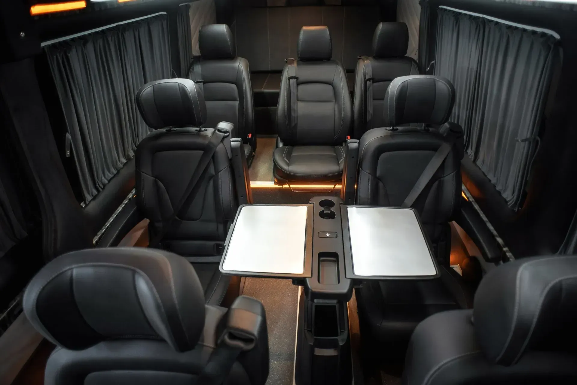 Interior of a luxury van with six black leather seats arranged around a table, curtains, and ambient lighting.