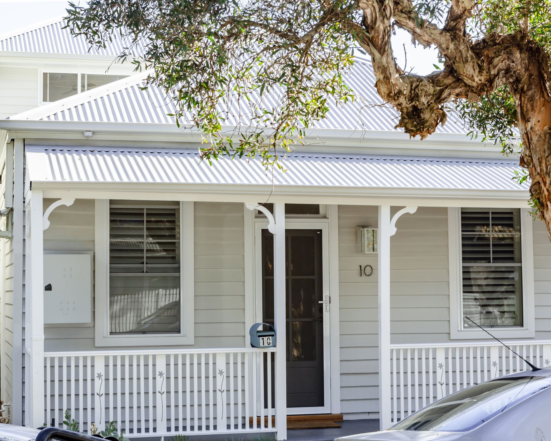 Small, light gray house with white trim, porch, and metal roof, with a tree branch overhead.