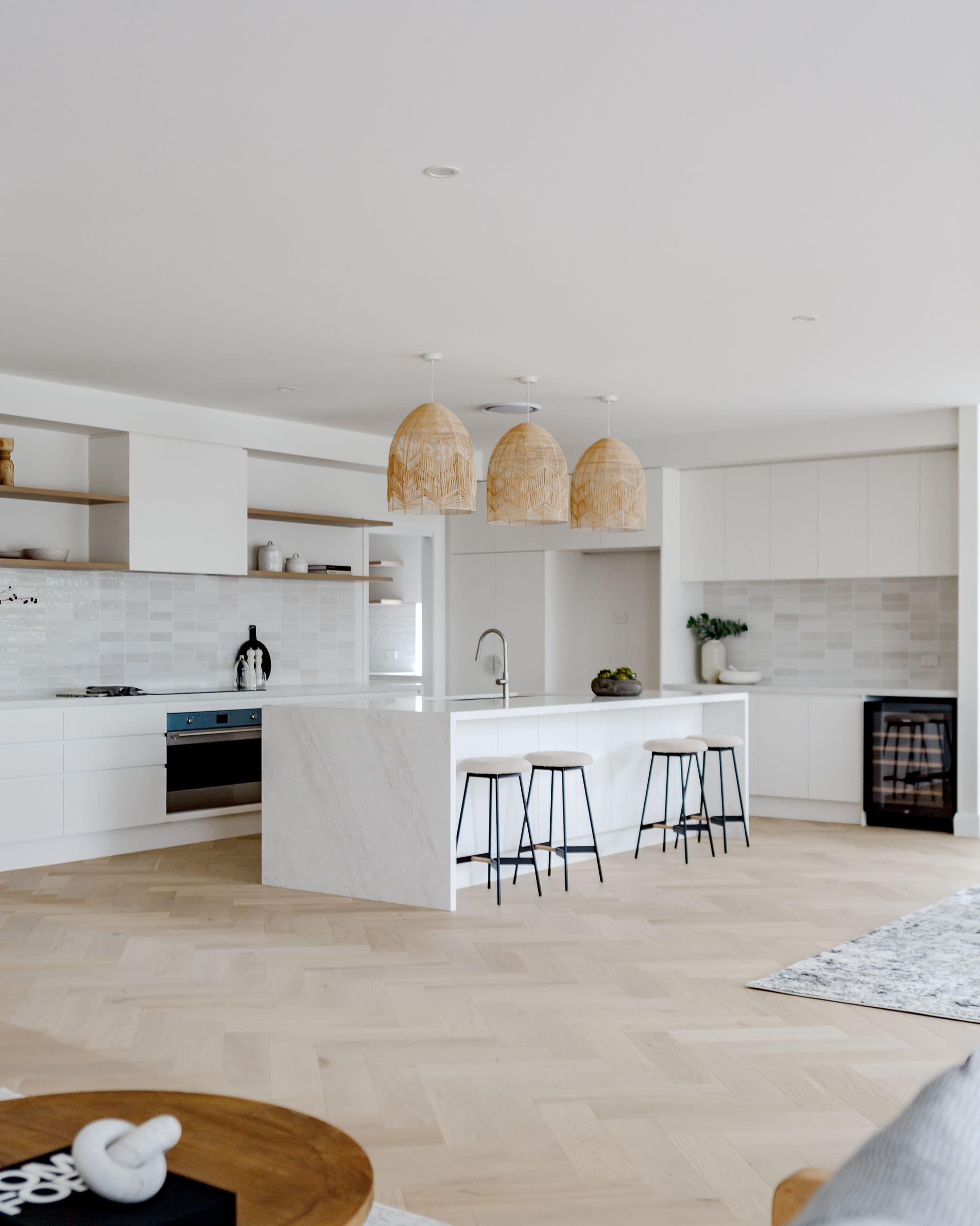 Modern white kitchen with island, wooden floors, wicker pendant lights, and stools.