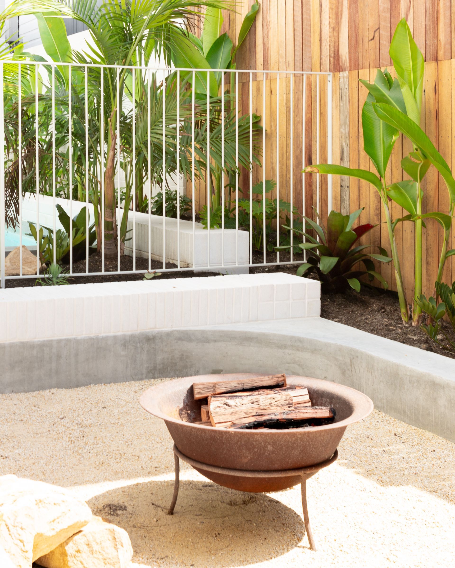 Outdoor fire pit with logs, sandy ground, white fence, wooden wall, and lush greenery.