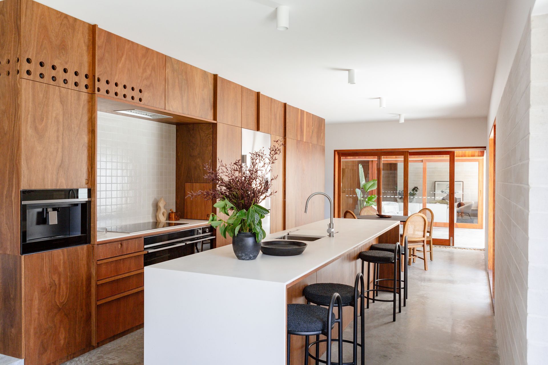 Modern kitchen with wood cabinets, white countertops, and an island with bar stools, leading to a patio.