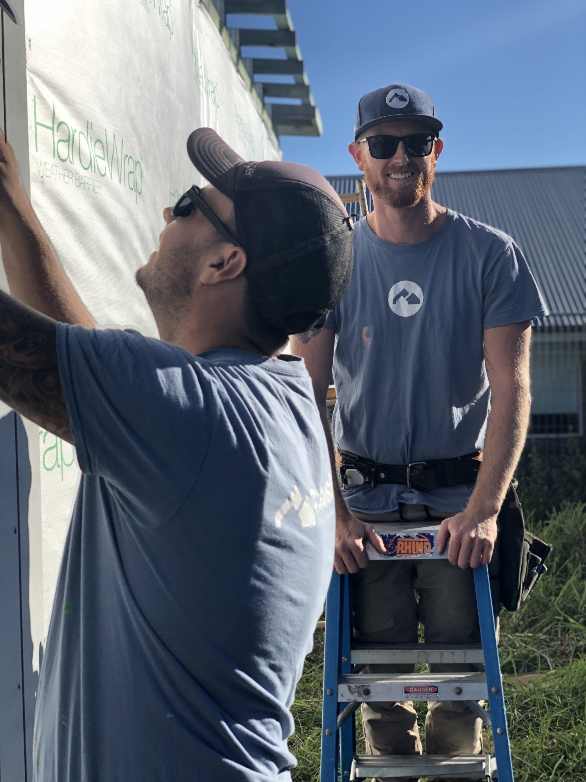 Two men, construction workers, installing siding on a building. One man on a ladder, the other holding the siding. Sunny day.