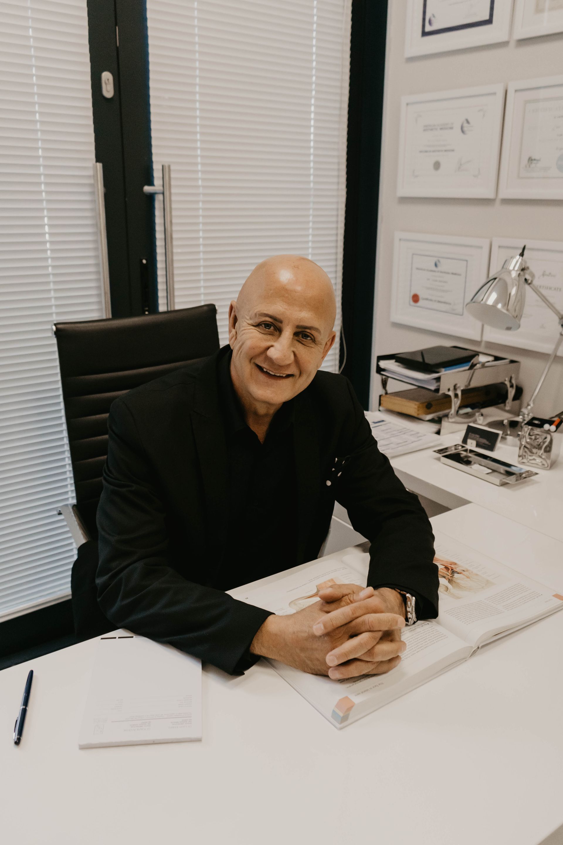 A man is sitting at a desk with his hands folded and smiling.