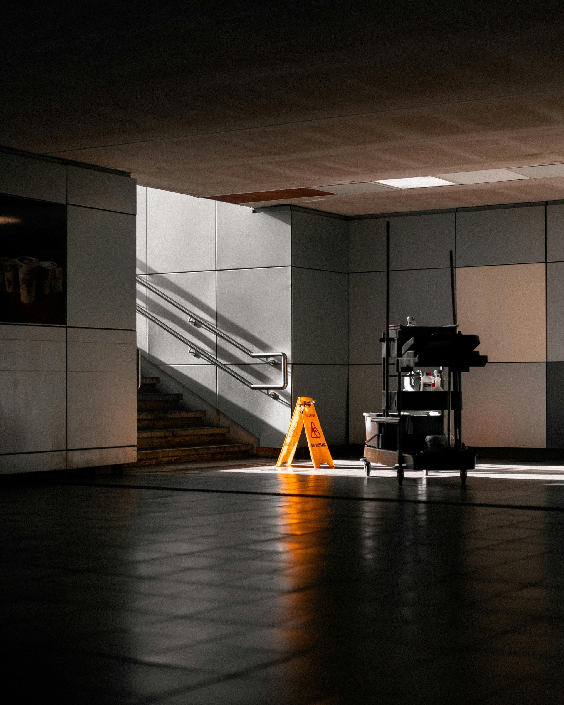 Dim airport concourse with an orange wet-floor sign near stairs and a maintenance cart in the background