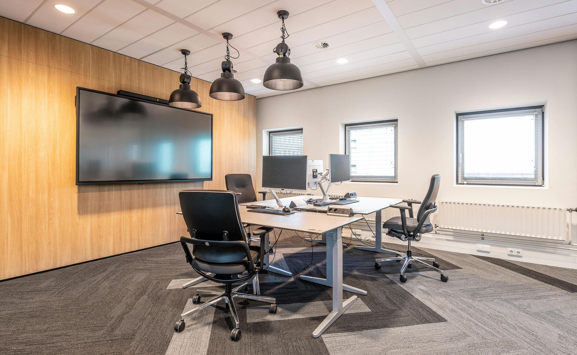 An office room with a large screen, wooden feature wall, modern desk, and black chairs on a grey patterned carpet.