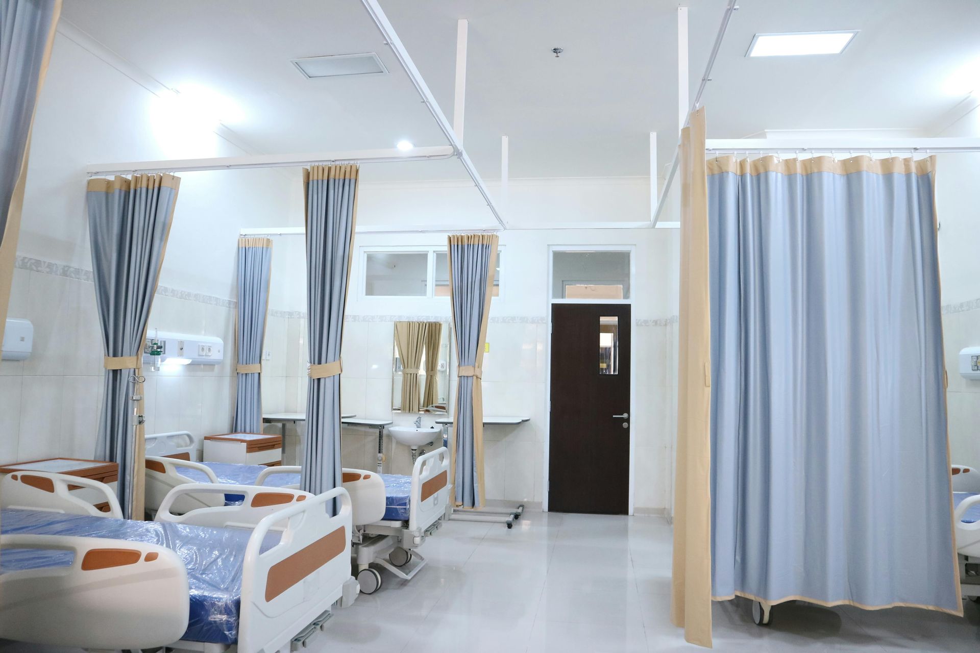 A hospital ward with several empty patient beds, gray privacy curtains, and a dark brown door in the background.