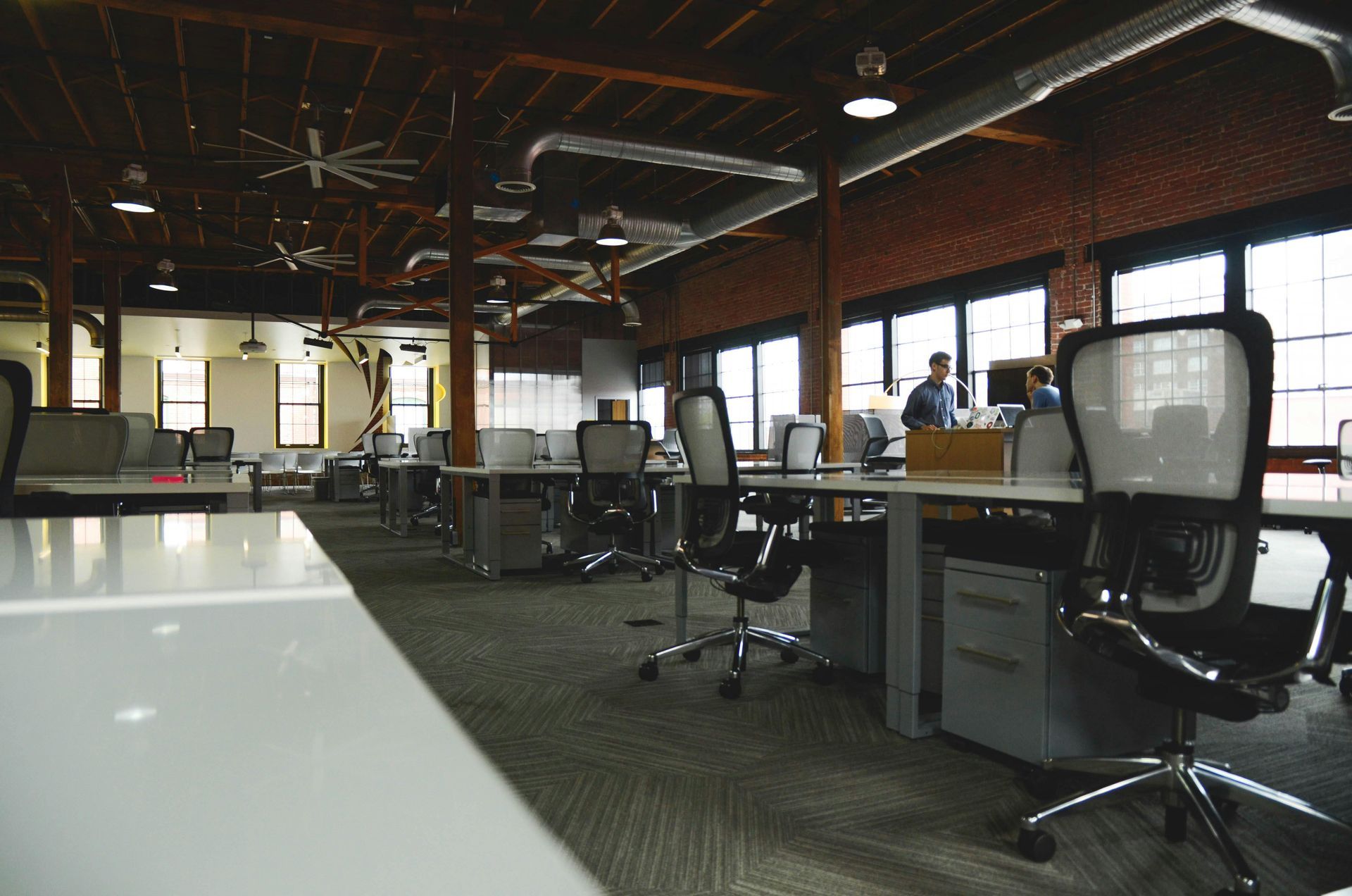 Modern open-plan office with rows of desks, rolling chairs, and exposed ceiling beams with industrial ductwork.