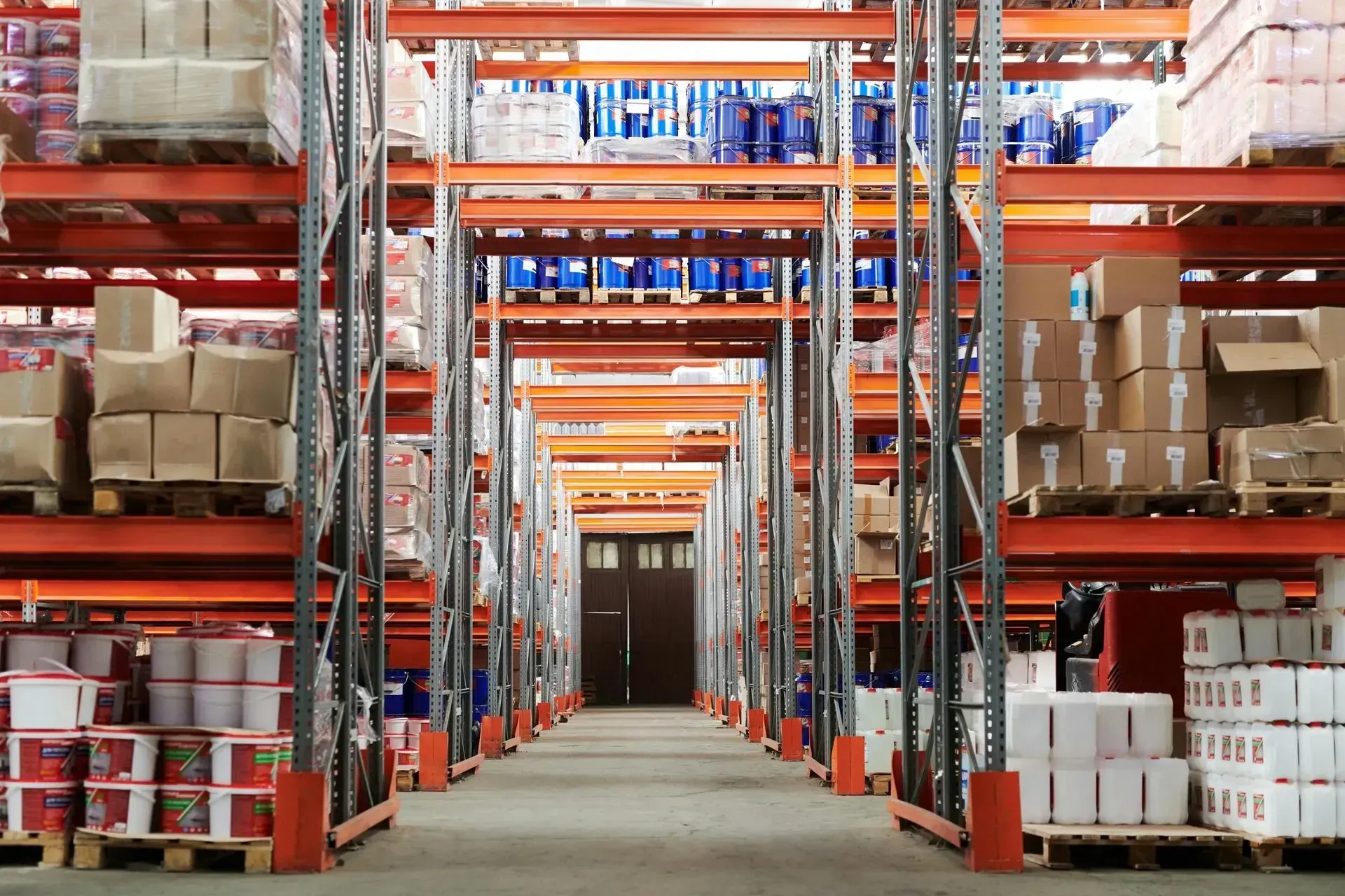 An aisle inside a warehouse with tall orange metal shelving units stacked with cardboard boxes and plastic drums.