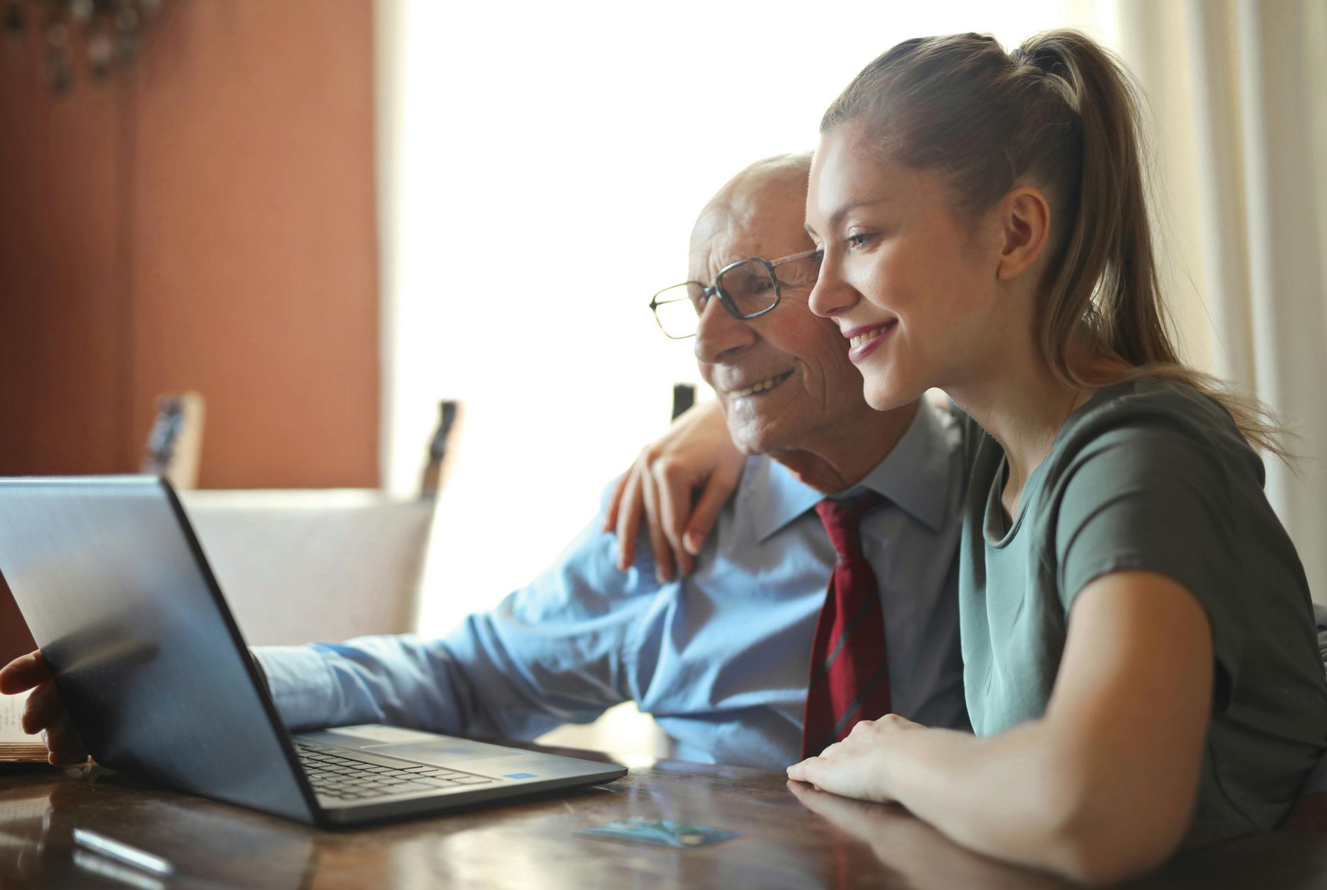 An elderly man and a young woman are sitting at a table looking at a laptop.