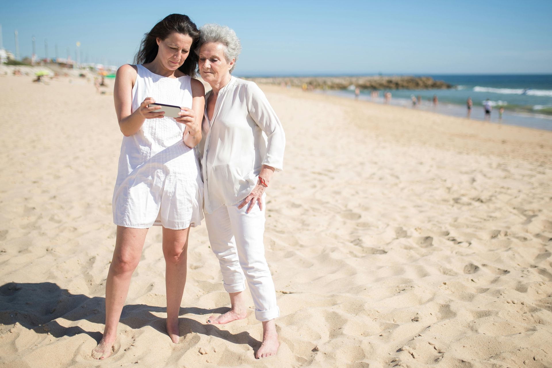 Two women are standing on a beach looking at a cell phone.