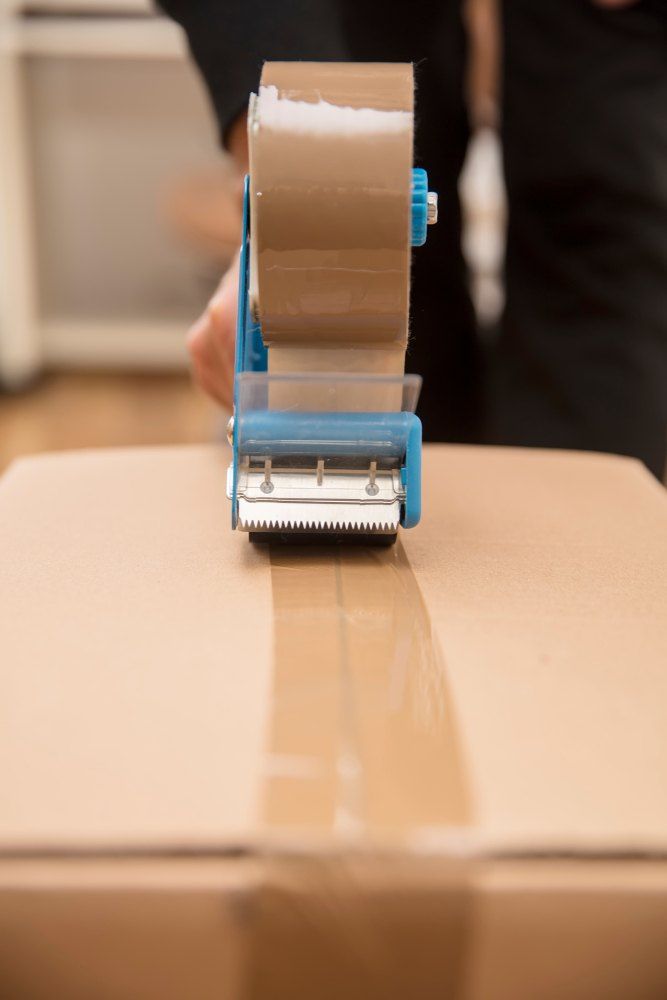 A Person is Using a Tape Dispenser to Tape a Cardboard Box — Buck Removals & Storage in Forbes, NSW