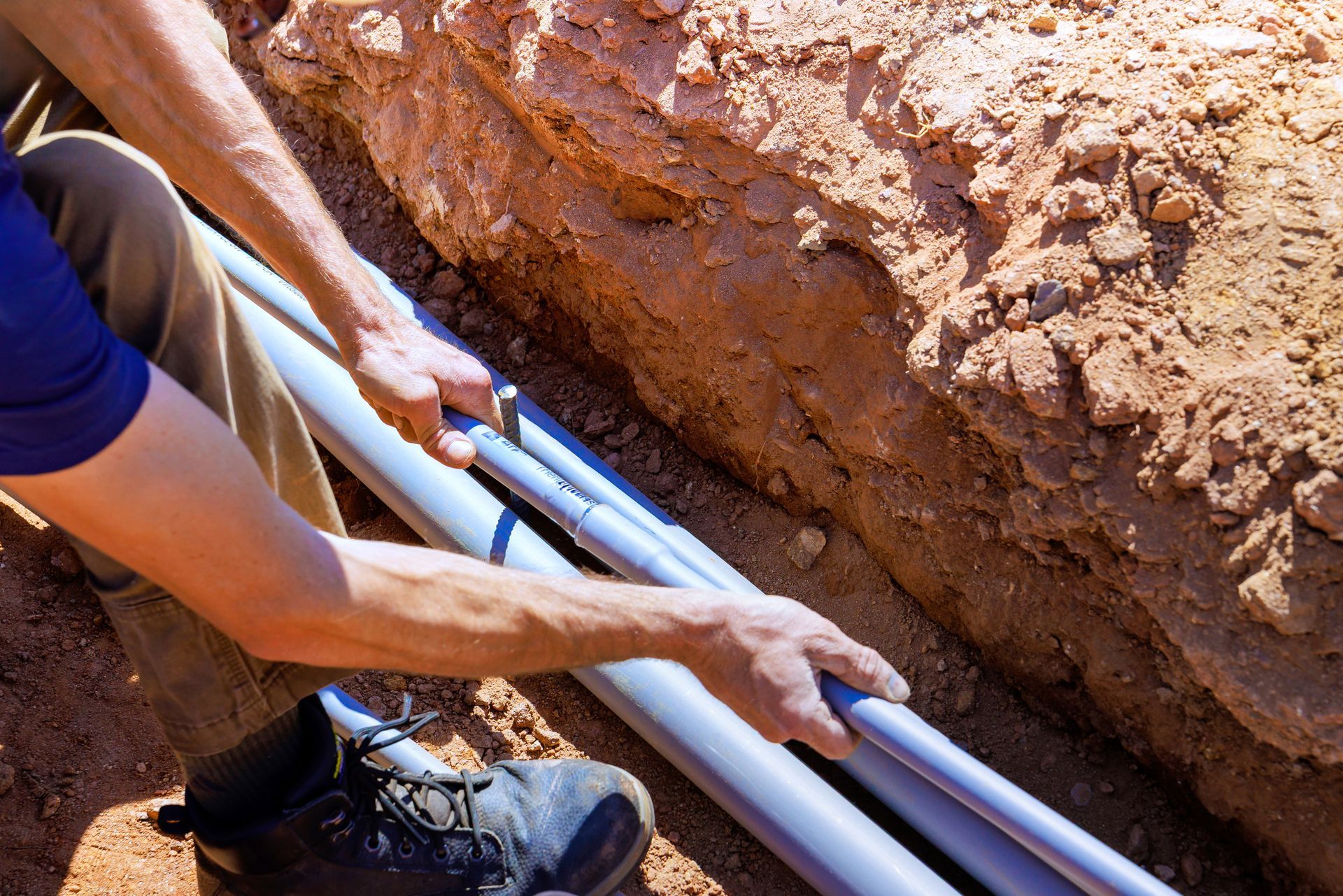 Worker laying gray pipes in a trench of red dirt