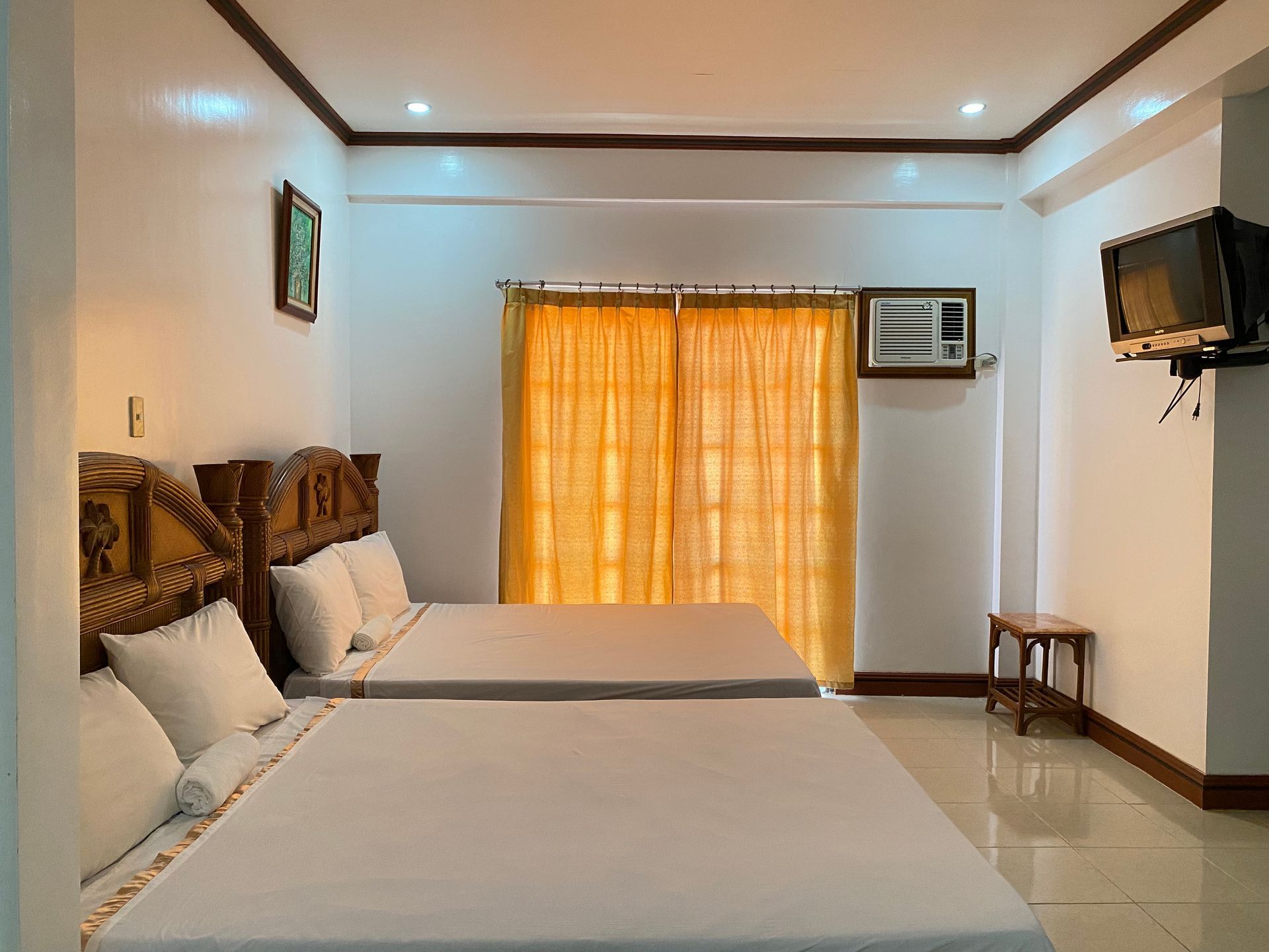 Bright hotel room with two beds, orange curtains, wall AC, and a TV on the right wall.