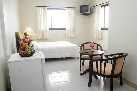 Small furnished bedroom with a bed, two chairs, a table, mini fridge, flowers, and wall-mounted TV