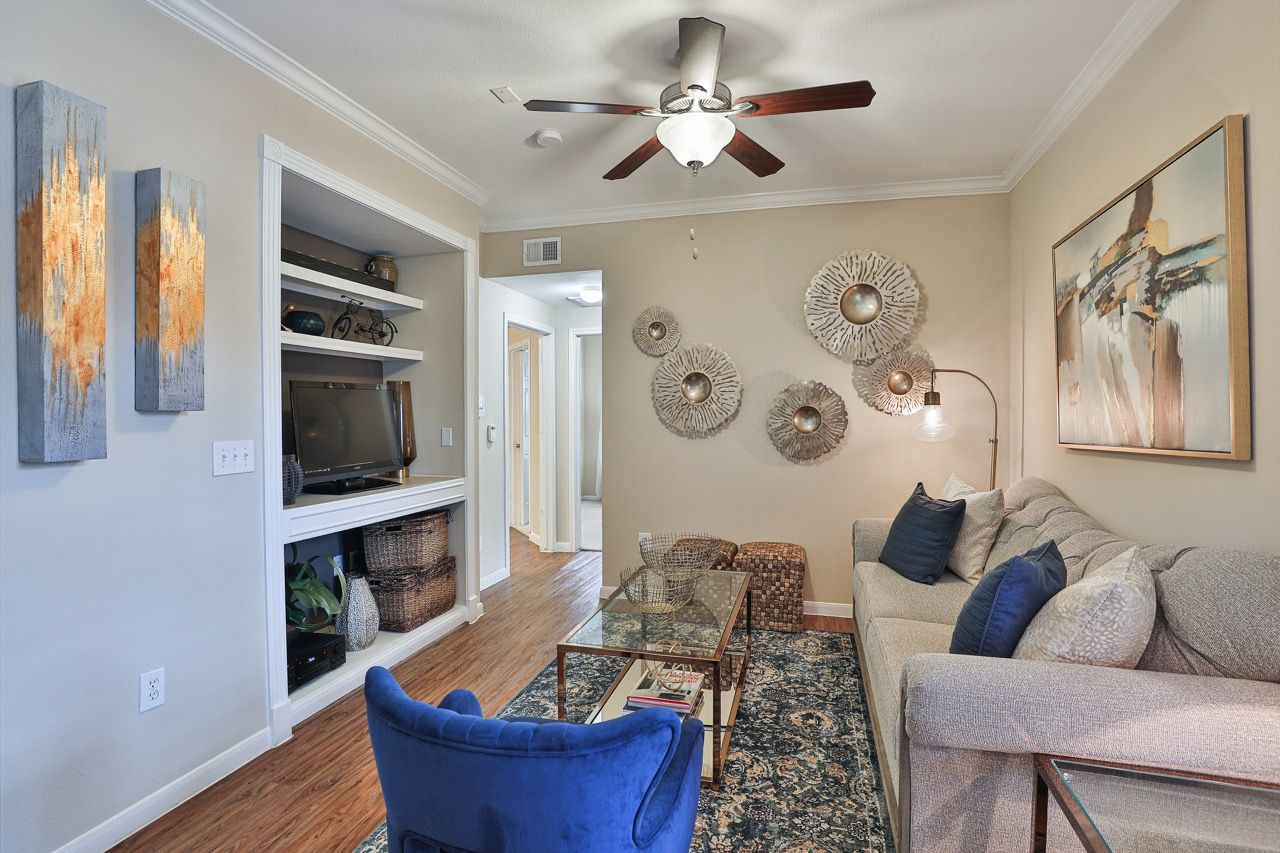 Living room in an apartment with beige walls, built-in shelves, TV, gray sofa, blue accent chair, and wall art.