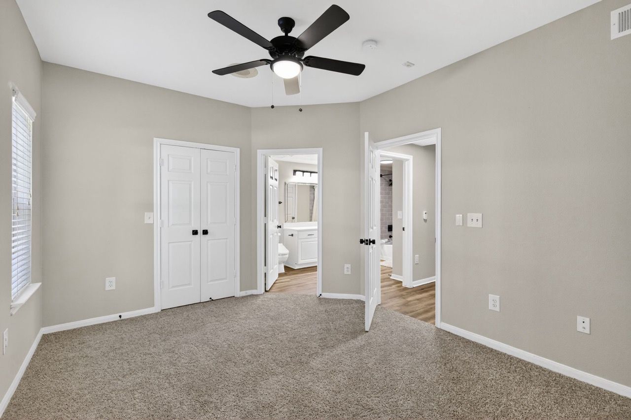 Interior of a neutral-toned bedroom with carpet, a ceiling fan, and doorway to a bathroom.
