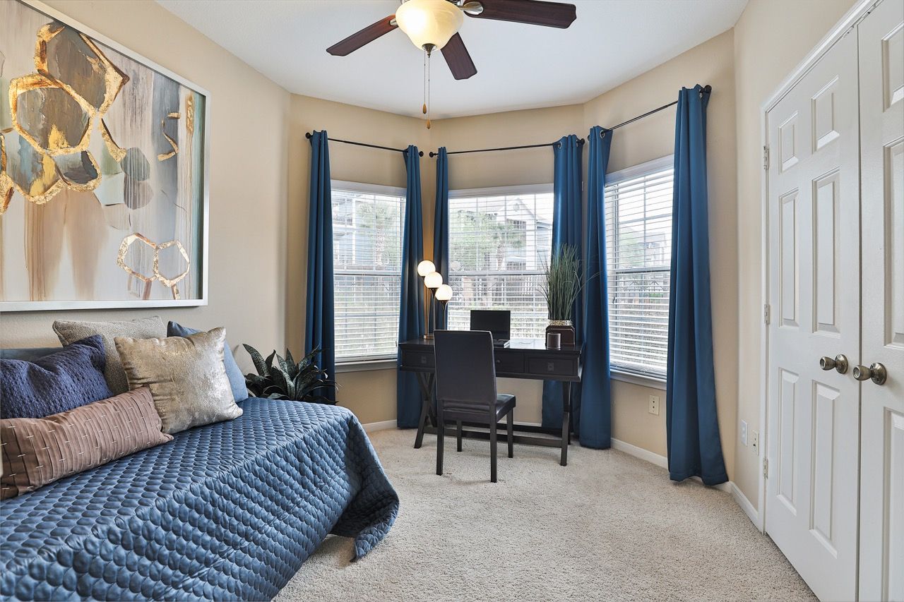 Bright apartment bedroom with bay windows, blue curtains, a desk, and a daybed.