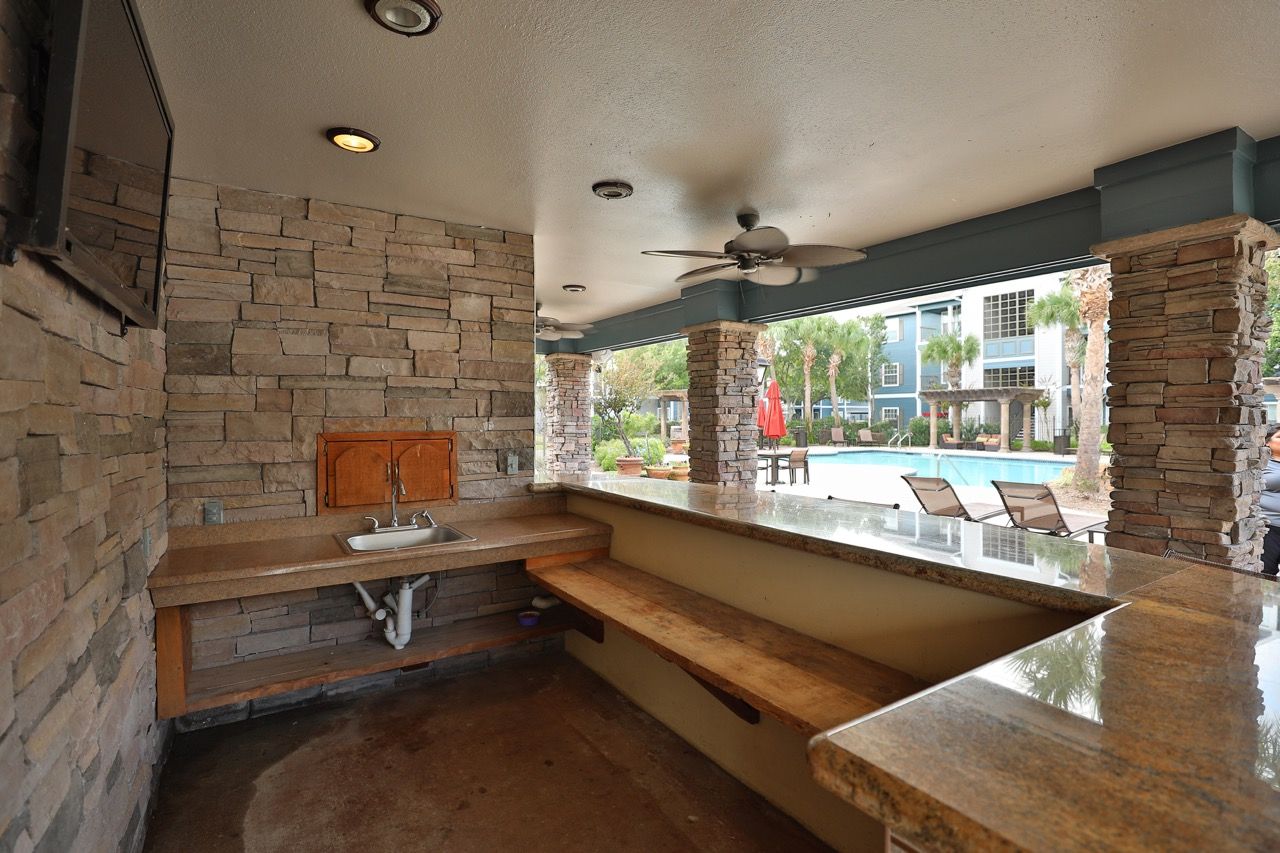 Outdoor communal grilling station with stone walls, sink, and granite counters near a pool.
