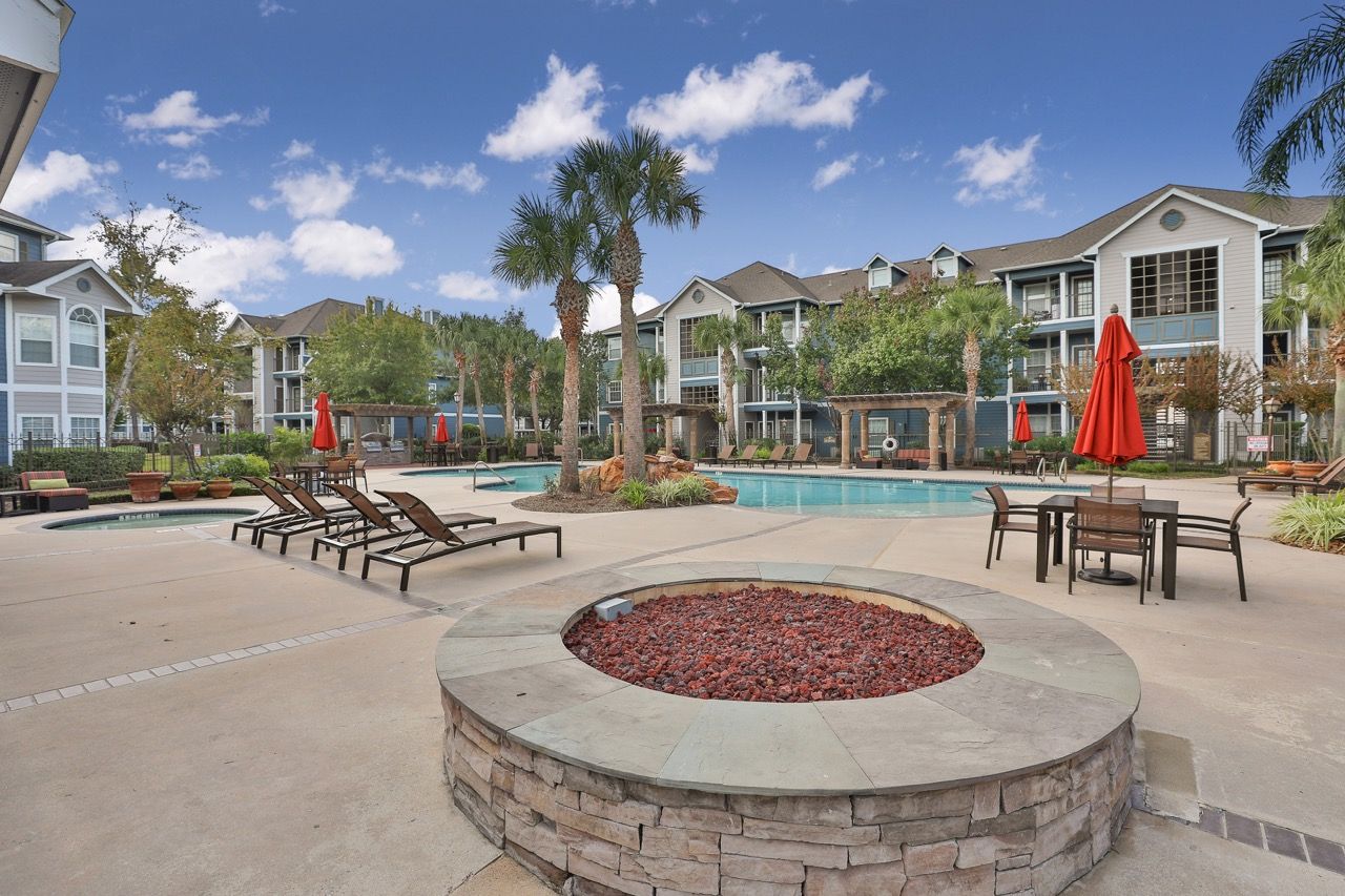 Outdoor apartment pool with lounge chairs, palm trees, and red umbrellas.