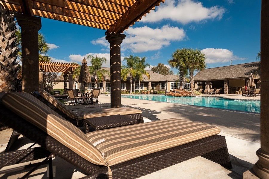 Lounge chairs on a shaded poolside patio with palm trees and a swimming pool under a sunny blue sky.