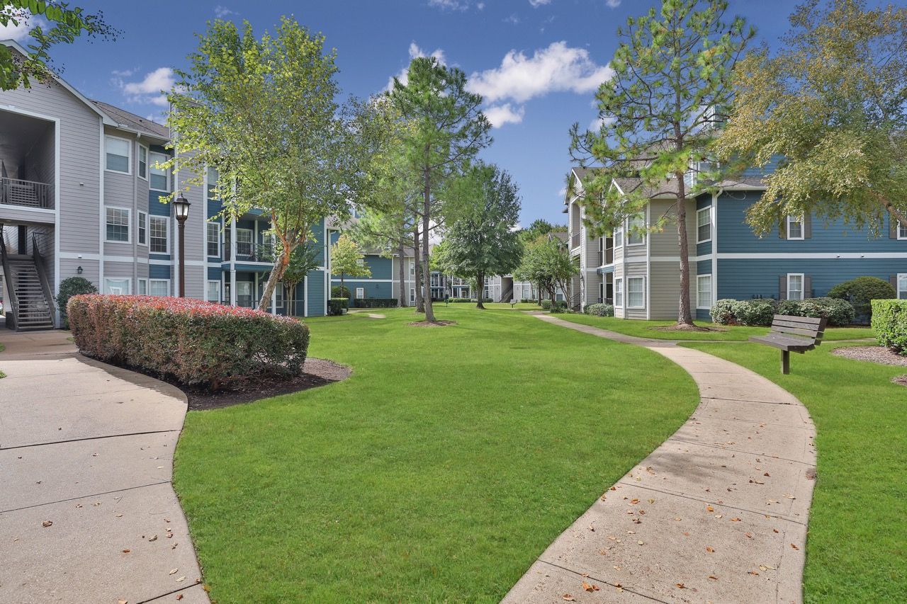 Curved concrete sidewalk through a green courtyard between blue apartment buildings and trees.