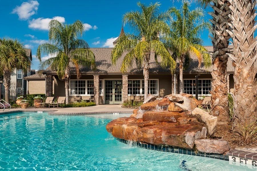 A resort-style swimming pool with a rock water feature, surrounded by palm trees and a clubhouse under a blue sky.