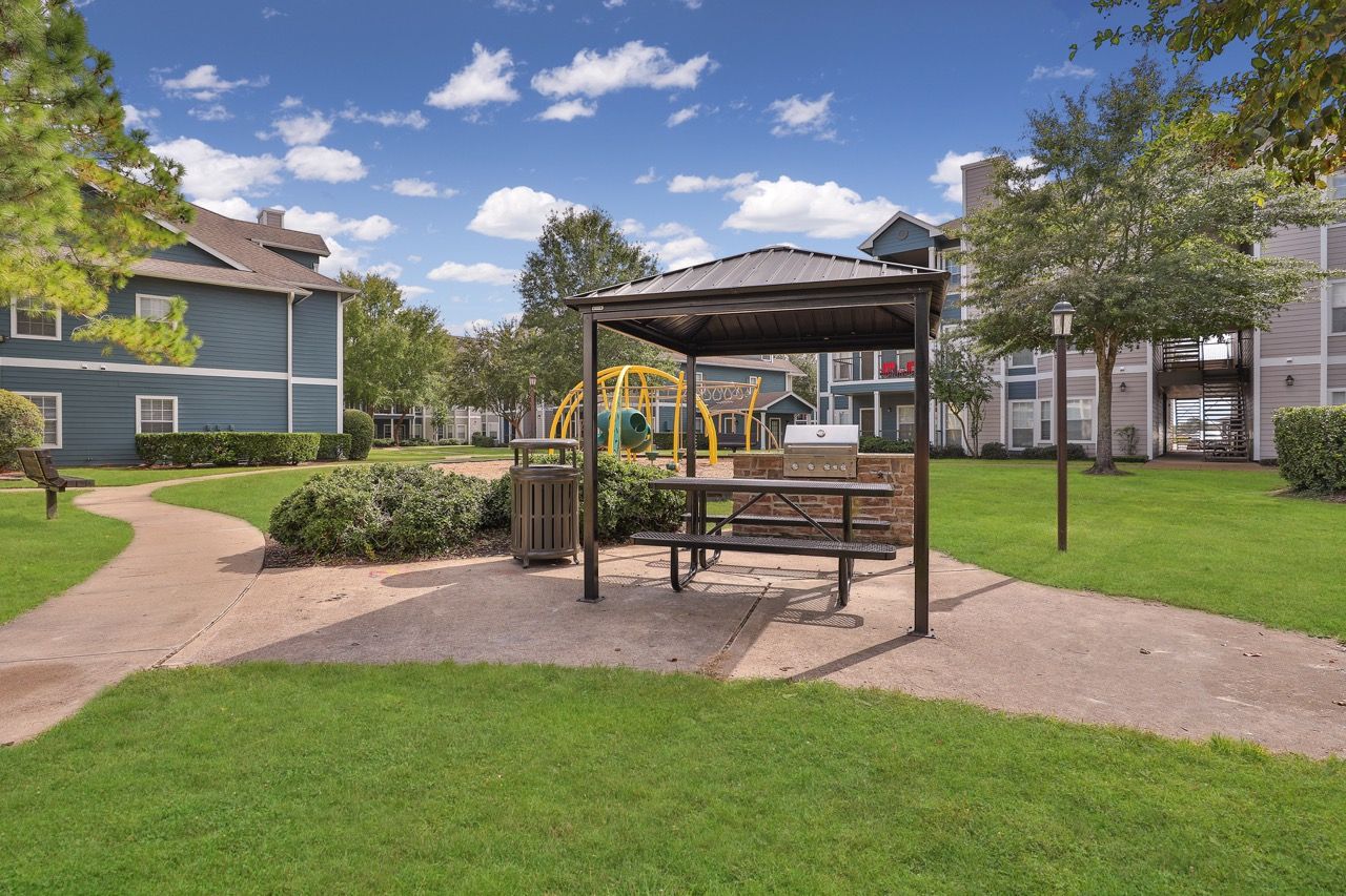Outdoor communal grilling area under a shelter with a picnic table, near a playground and green lawn.