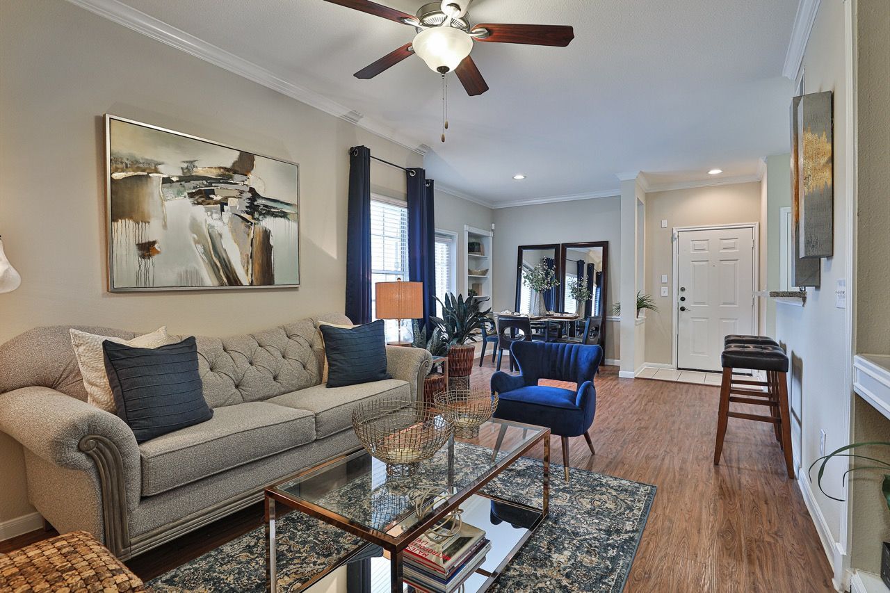 Living room in an apartment with a beige sofa, blue accent chair, glass coffee table, and dining area in the background.