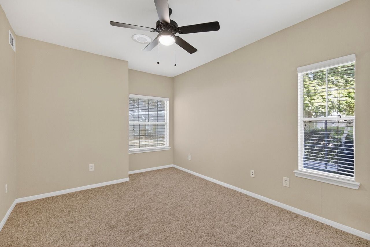 Interior bedroom with beige walls, carpet, two windows, and a ceiling fan.